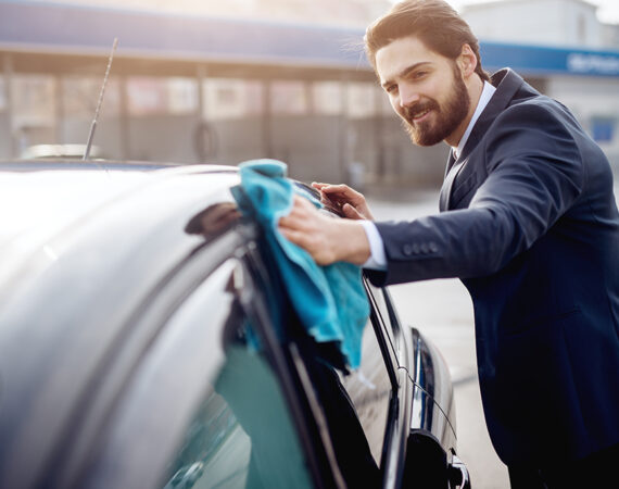 Young successful stylish bearded businessman in the suit cleaning the car with blue cloth outside.